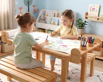 May include: A light-colored wooden activity table with a built-in paper roll dispenser and two integrated cylindrical pencil holders. The table features a smooth rectangular surface and is accompanied by a matching wooden bench for seating.
