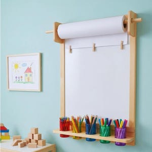 May include: A wall-mounted light wood paper roll dispenser and drawing board featuring a white paper sheet held by three wooden clips. A shelf below holds four colorful plastic cups filled with various colored pencils and markers.
