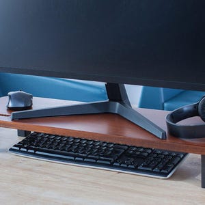 Op de afbeelding: Een houten monitorstandaard met zwarte metalen poten, die een computermonitor, toetsenbord en draadloze muis ondersteunt. Een koptelefoon ligt op de standaard.