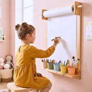 May include: A wooden art easel with a roll of white paper and a shelf holding colourful art supplies. A young person in a mustard-coloured dress is drawing on the paper with crayons. The easel is mounted on a pink wall.