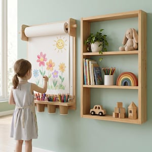 May include: A wall-mounted wooden paper roll dispenser with a drawing surface featuring a sun and flower illustration, accompanied by three attached cup holders for colored pencils, next to a three-tiered wooden wall shelf with toys.