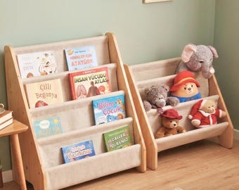 May include: Two light-colored wooden bookshelves with beige fabric slings. The left unit holds children's books with titles including Cilli Begonya and Bilim Çocuk. The right unit displays plush toys, including a gray elephant and teddy bears.
