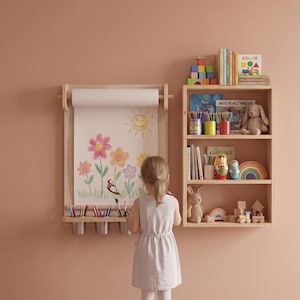 May include: A wall-mounted light wood paper roll holder with a drawing of a sun and flowers, featuring a shelf with three metal cups for art supplies, positioned next to a three-tiered wooden bookshelf filled with books, toys, and decor.