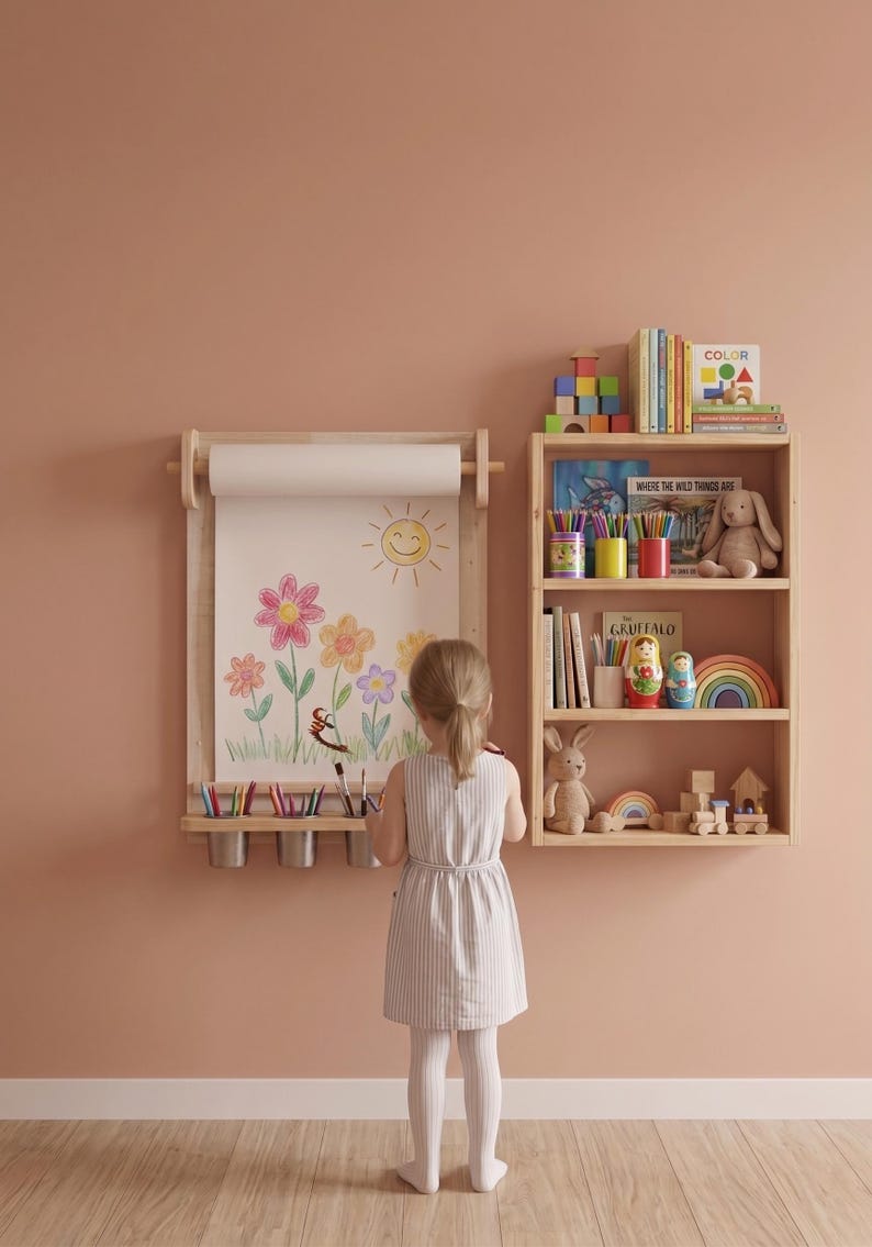 May include: A wall-mounted light wood paper roll holder with a drawing of a sun and flowers, featuring a shelf with three metal cups for art supplies, positioned next to a three-tiered wooden bookshelf filled with books, toys, and decor.
