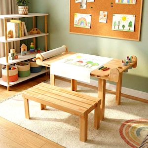 Könnte beinhalten: Ein Kindertisch aus hellem Holz mit Papierrolle, Bank und Stifthaltern. Der Tisch hat eine Zeichnung auf dem Papier. Eine Pinnwand mit Zeichnungen befindet sich an der Wand. Ein weißes Regal mit Spielzeug und Büchern befindet sich im Hintergrund.