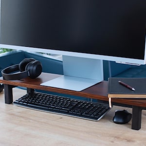 Puede incluir: Un soporte de monitor de madera con patas de metal negro, que sostiene un monitor de computadora, un teclado, unos auriculares, un teléfono y un cuaderno con un bolígrafo.