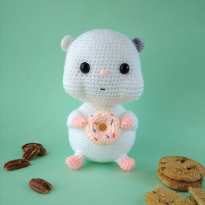 May include: A white crocheted hamster doll holding a pink donut with colorful sprinkles. The hamster has black eyes, a pink nose, and gray ears. Pecans and cookies are scattered around the doll on a light green background.