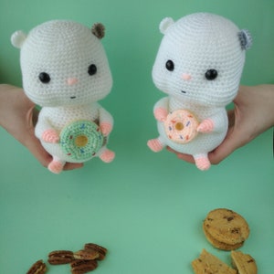 May include: Two white crochet hamster dolls, each holding a donut. One donut is green with sprinkles, the other is pink. The hamsters have black eyes, pink noses, and small ears. Pecans and cookies are scattered below.