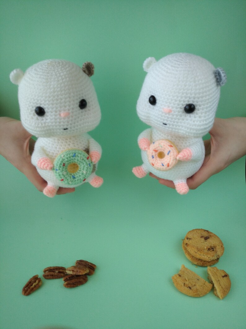May include: Two white crochet hamster dolls, each holding a donut. One donut is green with sprinkles, the other is pink. The hamsters have black eyes, pink noses, and small ears. Pecans and cookies are scattered below.