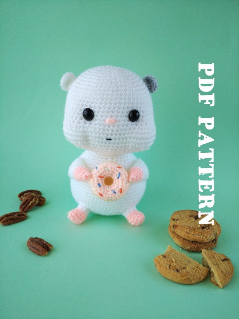 May include: A white crocheted hamster doll holding a donut, with black eyes, pink nose and feet, and gray ears. The doll is on a light green background with pecans and cookies. Text on the right says "PDF PATTERN".