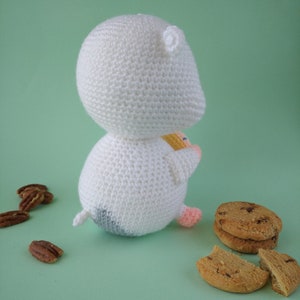 May include: A handmade white crochet hamster toy with a small tail, black and gray bottom, and pink feet. The hamster is next to chocolate chip cookies and pecans on a light green surface.