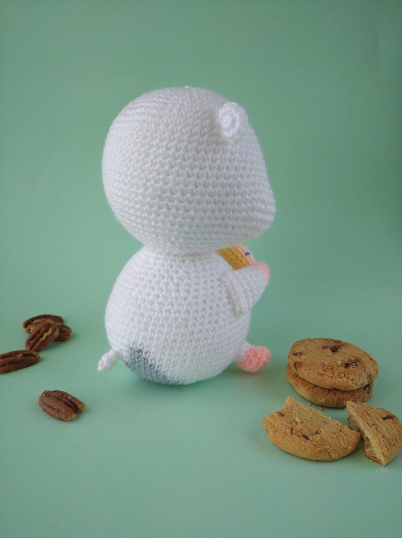 May include: A handmade white crochet hamster toy with a small tail, black and gray bottom, and pink feet. The hamster is next to chocolate chip cookies and pecans on a light green surface.