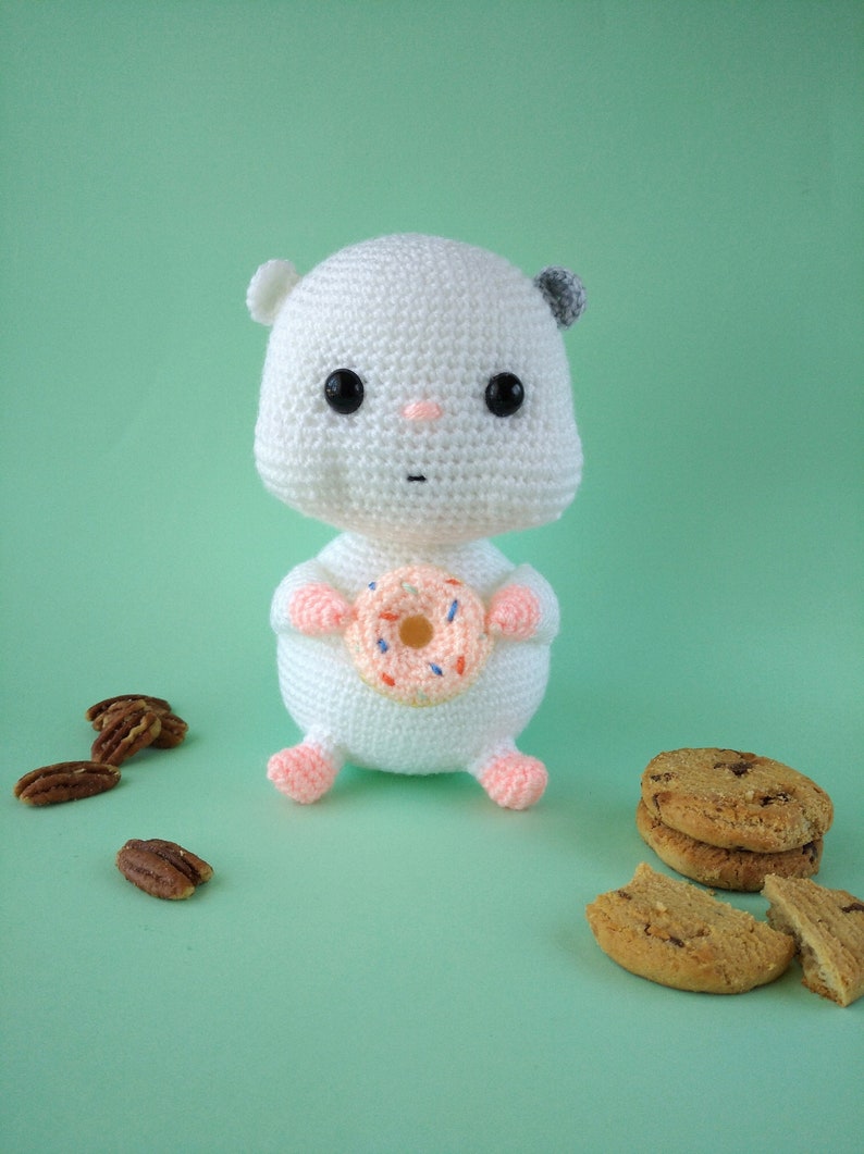 May include: A white crocheted hamster doll holding a pink donut with colorful sprinkles. The hamster has black eyes, a pink nose, and gray ears. Pecans and cookies are scattered around the doll on a light green background.