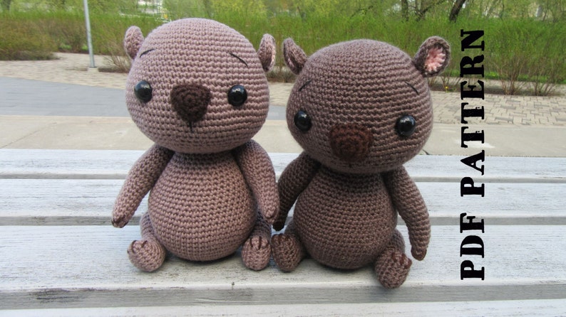 May include: Two brown crocheted stuffed animals with black eyes and brown noses. The animals are sitting on a wooden bench. The text "PDF PATTERN" is visible in the background.