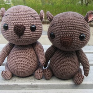 May include: Two brown crocheted stuffed animals sitting on a wooden bench. The animals have black eyes, brown noses, and pink ears.