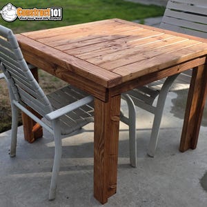 May include: A square wooden table with a natural brown finish and visible wood grain, featuring a slatted top design and four sturdy legs. The table is accompanied by two light gray metal chairs with horizontal slats on the backrests.
