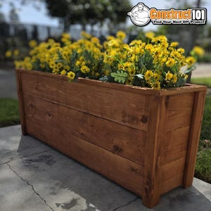 May include: A rectangular wooden planter box with a medium brown stain and visible wood grain, filled with a dense arrangement of bright yellow flowers. The planter features horizontal wooden planks and vertical corner supports.