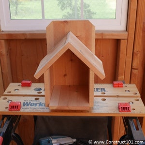 May include: A wooden birdhouse with a simple design, featuring a pitched roof and a rectangular opening. The birdhouse is unfinished and appears to be made of cedar wood. It is sitting on a workbench with a ruler and clamps.