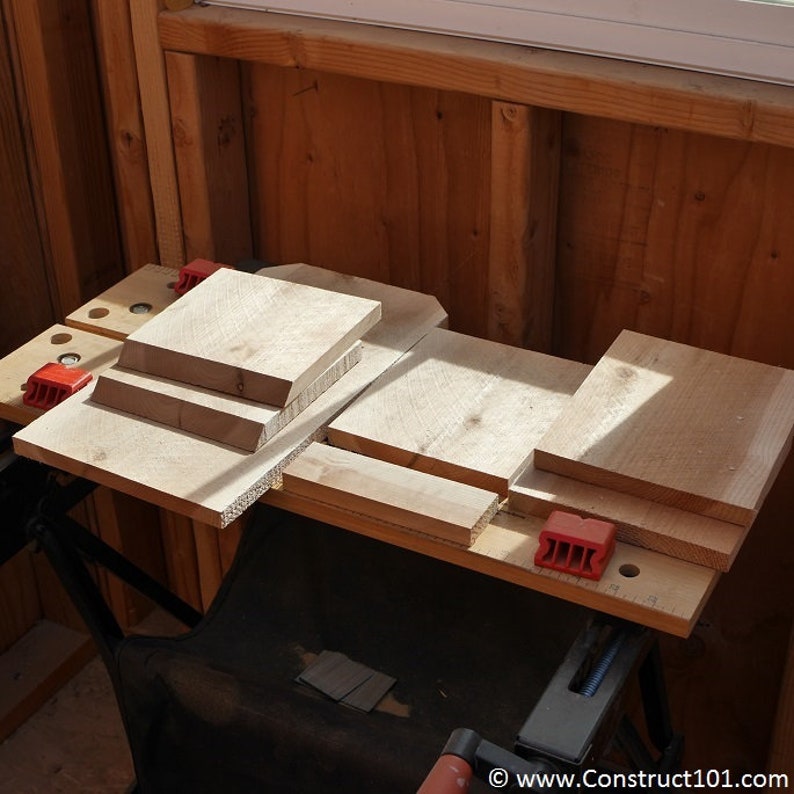 May include: A close-up of a workbench with several pieces of wood cut to different sizes. The wood is arranged in a haphazard manner, and there are red clamps holding the pieces together. The workbench is made of wood and has a black metal frame.