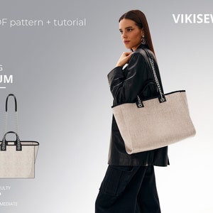 Può includere: Una borsa tote beige con un bordo nero e una tracolla a catena argentata. La borsa è tenuta da una persona che indossa una giacca di pelle nera e jeans. Il testo "VIKISEWS" è visibile nell'angolo in alto a destra dell'immagine. Il testo "BAG KUM" è visibile nell'angolo in alto a sinistra dell'immagine.