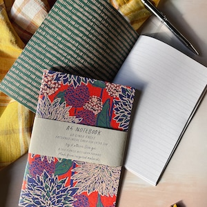 May include: A5 notebook with a floral design on a red background, with a paper band that reads "A5 Notebook". A green striped notebook and a lined notebook are also visible, along with a silver pen. The background includes a yellow plaid cloth.