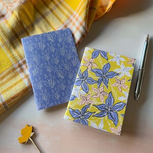 May include: Two small notebooks with floral patterns. One notebook has a blue cover with a white floral pattern. The other notebook has a yellow cover with a blue and pink floral pattern.