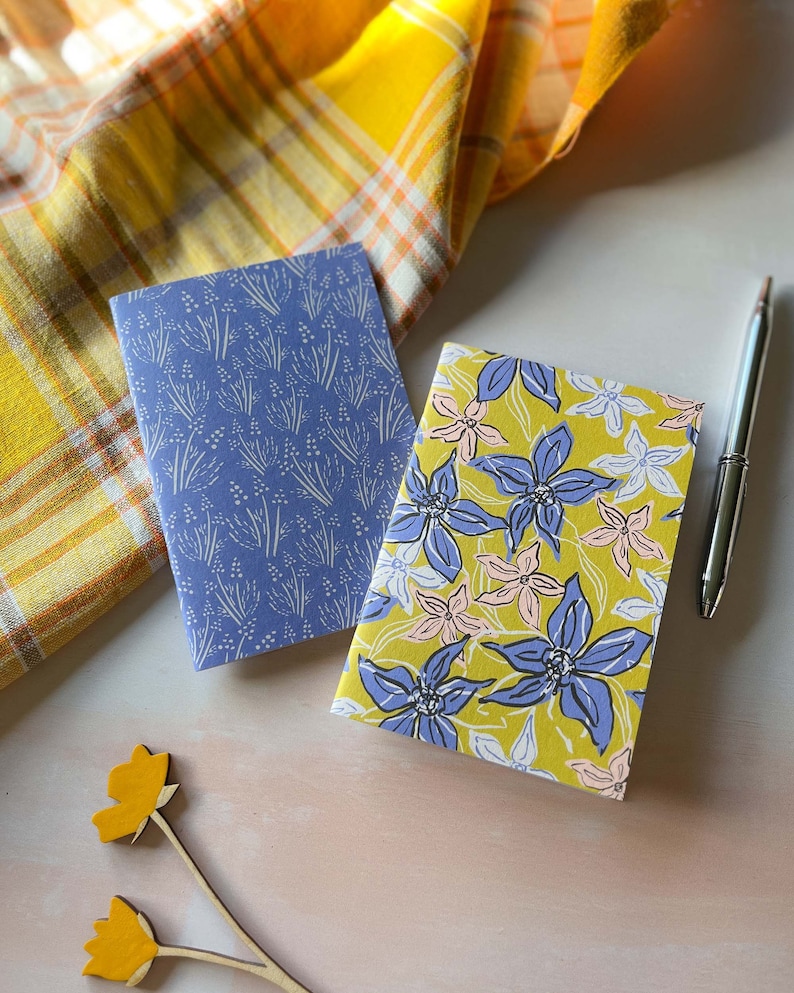 May include: Two small notebooks with floral patterns. One notebook has a blue cover with a white floral pattern. The other notebook has a yellow cover with a blue and pink floral pattern.