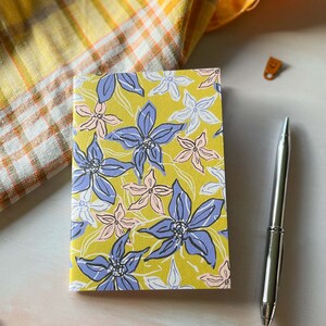 May include: A yellow notebook with a floral pattern featuring blue, pink, and white flowers. The notebook is closed and lying on a white surface.