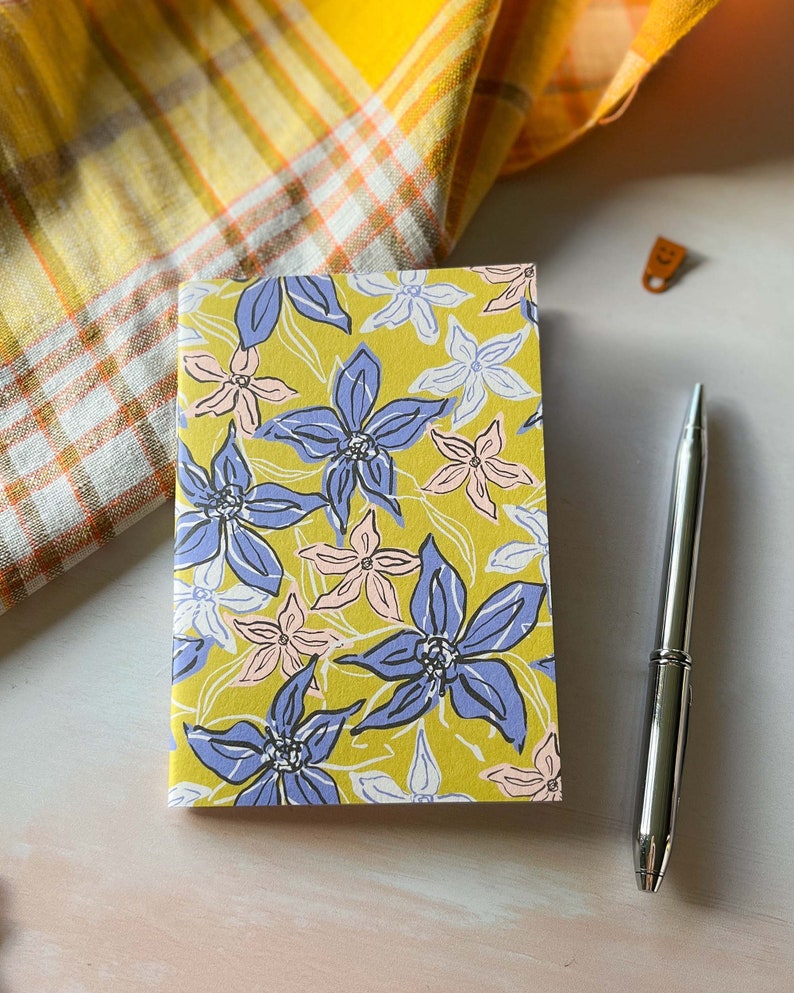 May include: A yellow notebook with a floral pattern featuring blue, pink, and white flowers. The notebook is closed and lying on a white surface.