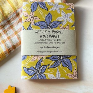 May include: Set of two pocket notebooks with a floral design on the cover. The notebooks are A6 size and have 40 plain pages each. The inside cover is patterned and the notebooks are made from recycled materials. "SET OF 2 POCKET NOTEBOOKS 40 PLAIN PAGES I A6 SIZE PATTERNED INSIDE COVER FOR EXTRA JOY by Kattern Design Made from recycled materials"