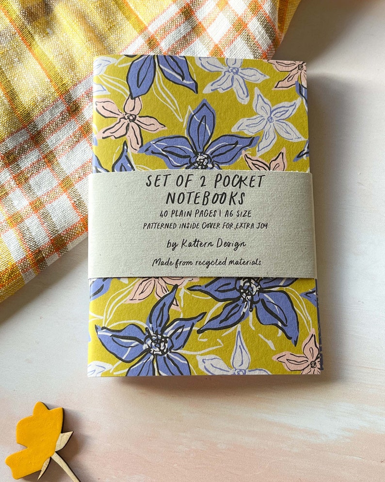 May include: Set of two pocket notebooks with a floral design on the cover. The notebooks are A6 size and have 40 plain pages each. The inside cover is patterned and the notebooks are made from recycled materials. "SET OF 2 POCKET NOTEBOOKS 40 PLAIN PAGES I A6 SIZE PATTERNED INSIDE COVER FOR EXTRA JOY by Kattern Design Made from recycled materials"