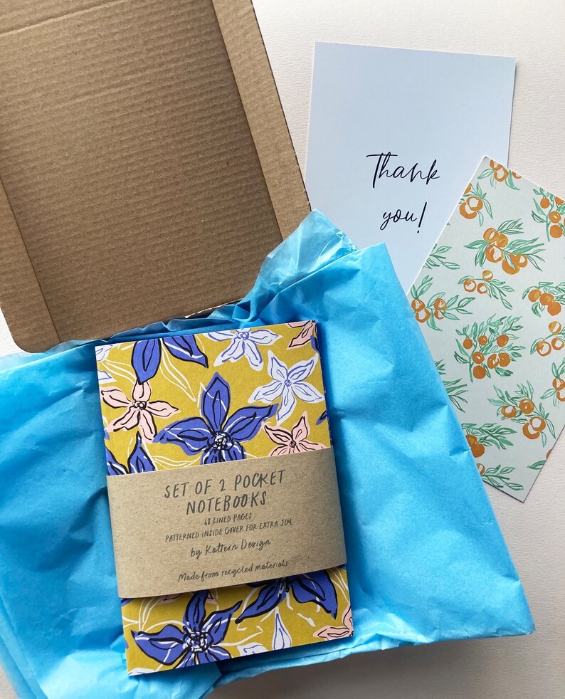 May include: Set of two pocket notebooks with floral designs on the covers. The notebooks are lined and have a patterned inside cover. The notebooks are made from recycled materials. The text on the cover reads "Set of 2 Pocket Notebooks 43 Lined Pages Patterned Inside Cover for Extra Joy by Kattern Design Made from recycled materials".
