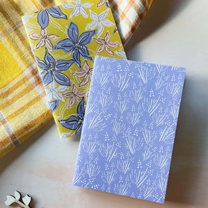 May include: Two small notebooks with floral patterns. One notebook has a yellow background with blue and pink flowers. The other notebook has a light blue background with white flowers.