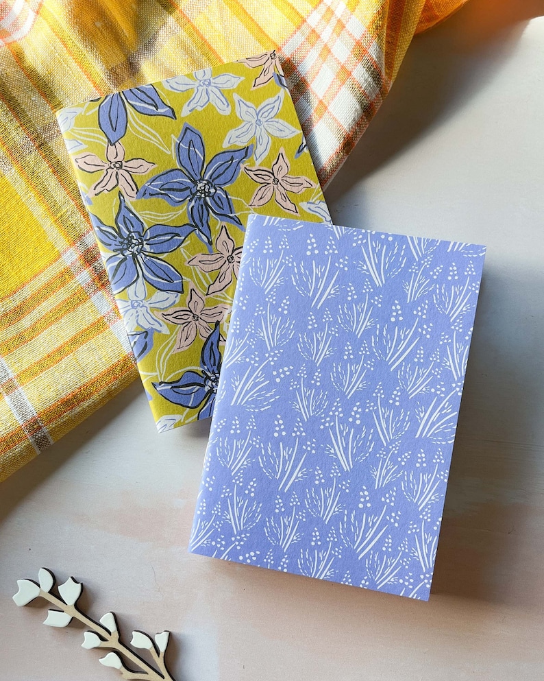 May include: Two small notebooks with floral patterns. One notebook has a yellow background with blue and pink flowers. The other notebook has a light blue background with white flowers.