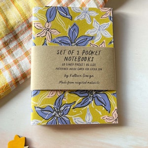 May include: Set of two pocket notebooks with a floral design on the cover. The notebooks are made from recycled materials and have 40 lined pages each. The cover is a yellow colour with blue, pink, and white flowers.