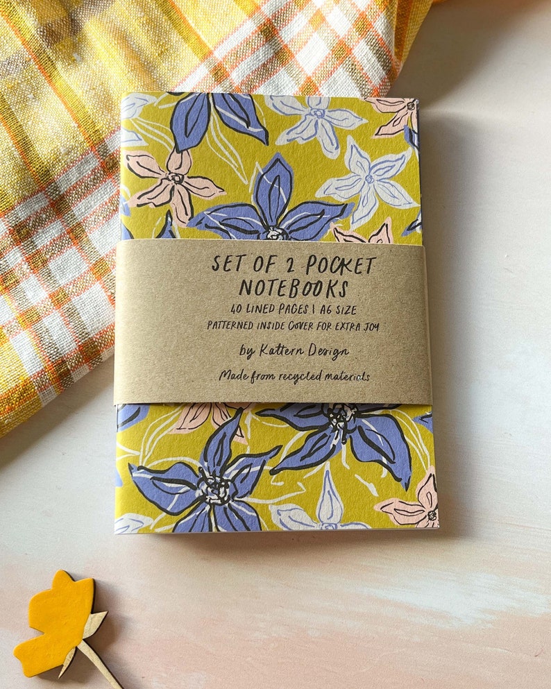 May include: Set of two pocket notebooks with a floral design on the cover. The notebooks are made from recycled materials and have 40 lined pages each. The cover is a yellow colour with blue, pink, and white flowers.