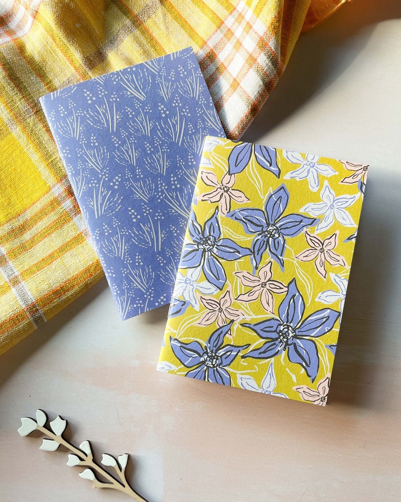 May include: Two small notebooks with floral patterns. One notebook has a blue background with white floral designs. The other notebook has a yellow background with blue and pink floral designs.
