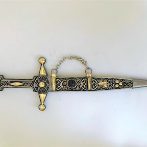 May include: A silver and gold dagger with a black handle and intricate gold filigree designs. The dagger has a chain attached to the handle.