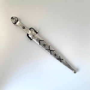 May include: A silver dagger with intricate floral designs and black accents. The dagger has a pointed tip and a decorative handle.