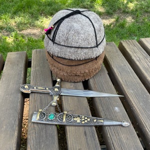 Puede incluir: Un sombrero de fieltro gris y marrón con un borla negra se encuentra sobre un banco de madera junto a una daga de plata con mango de oro y una piedra preciosa verde.