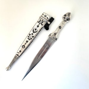 May include: A silver dagger with a black and silver patterned handle and a matching sheath. The dagger has a sharp point and a curved blade.