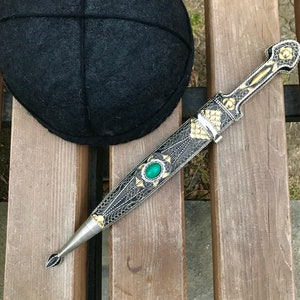 May include: An ornate dagger with a silver, gold, and black design, featuring a green gemstone in the center. The dagger is in its sheath, and a black hat is in the background. The dagger's sheath is approximately 10 inches long.