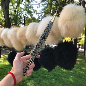 May include: A decorative dagger with intricate gold and black designs and green gemstones. The dagger is held in a hand with a red bracelet.