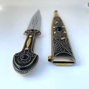 以下が含まれることがあります： 黒のハンドルと装飾的な鞘が付いた銀と金の短剣。ハンドルは複雑なフィリグリーと黒い宝石で飾られています。