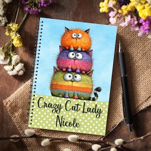 May include: Spiral-bound notebook with a design of three cartoon cats in orange, purple, and green. The notebook has a light blue background and a green polka dot border with the text "Crazy Cat Lady Nicole."
