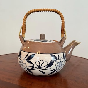 May include: A ceramic teapot with a brown top and a cream-colored base decorated with blue floral designs. It features a woven handle and a spout with a gold-colored trim. The lid has a small brown knob.