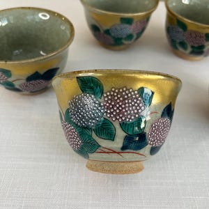 May include: Set of five small ceramic teacups with a gold rim and floral design. The cups feature a white background with green leaves and pink and white flowers.