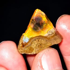 Könnte beinhalten: Ein roher Opal-Edelstein in dreieckiger Form mit lebendigen Farben. Der Opal zeigt eine Mischung aus Gelb, Orange und Hauch von Blau und Lila. Die Basis des Steins ist eine strukturierte, hellbraune Farbe, gehalten von Fingern.