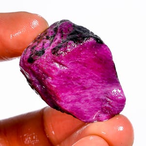 Peut inclure: Une pierre précieuse de rubis naturelle, brute, d'un magenta vif. Le rubis a une surface texturée avec des inclusions noires foncées et est tenu entre les doigts sur un fond blanc. La pierre mesure environ 3.8 cm de long.
