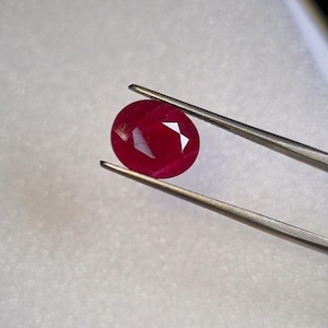 May include: A vibrant, deep red oval-cut gemstone held by a pair of silver-toned metal tweezers. The stone features a faceted surface that reflects light, showcasing its rich crimson color and translucent quality against a textured white background.